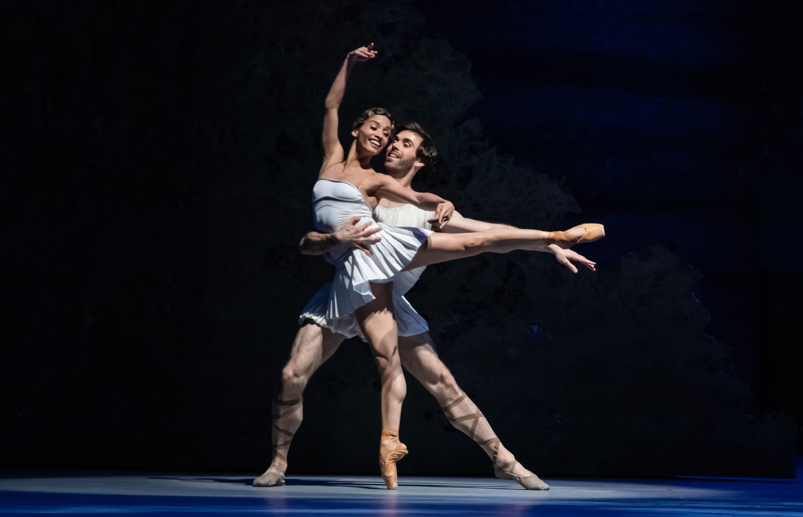 Karina Gonzalez performs a piqué arabesque on her right leg with her right arm high and her left arm held out to the side. She smiles joyfully and looks over her left shoulder towards her partner, Connor Walsh, who holds onto her waist while lunging behind her. Her costume is a short, blue dance dress and tan pointe shoes, while he wears a white toga and brown ballet shoes.