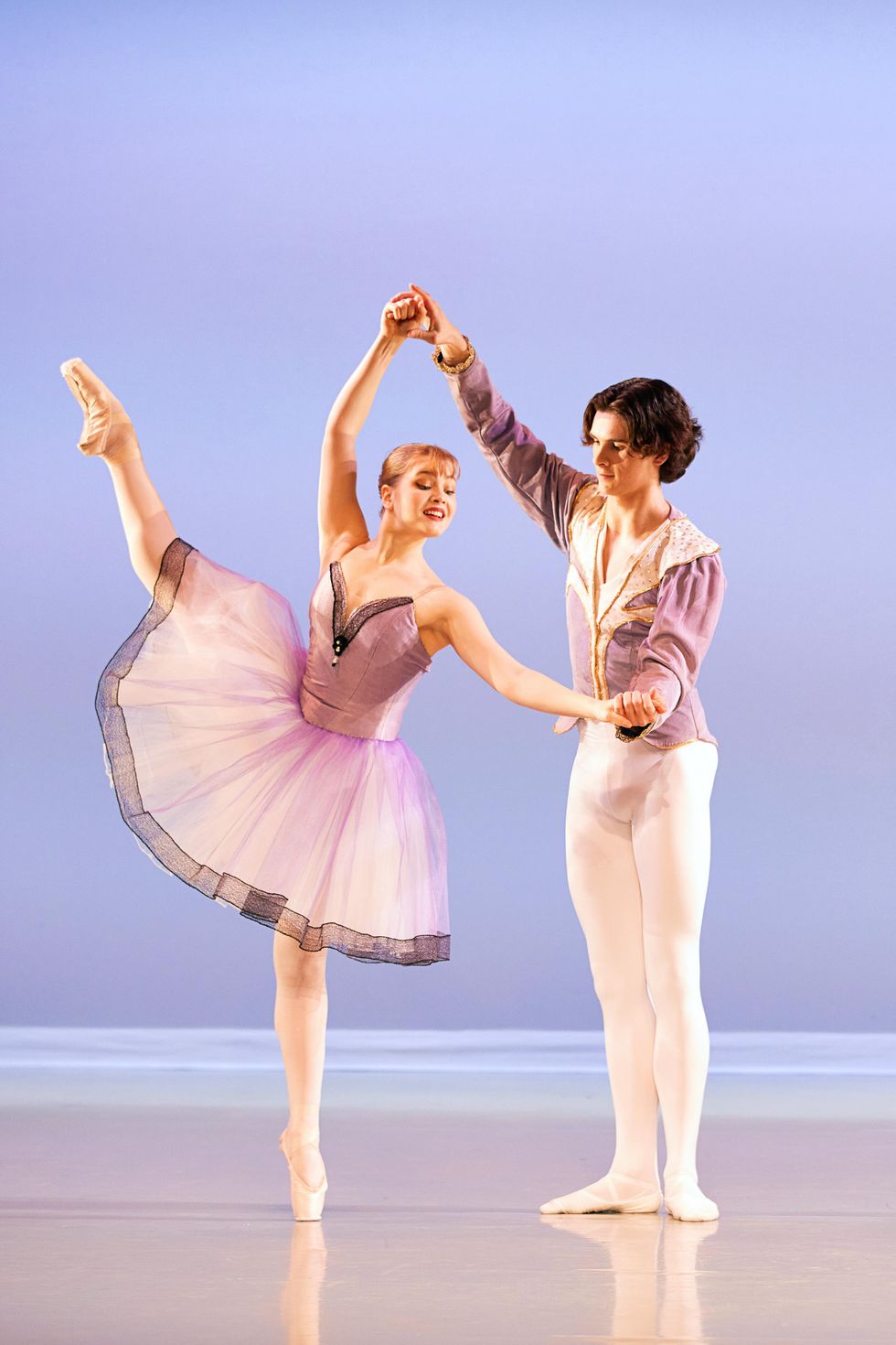 A redheaded teenage ballerina in a light purple tutu does a du00e9veloppu00e9 ecartu00e9 derriere with her right leg and stands on pointe. Her male partner, in white tights and a light purple tunic, holds her left hand to the side and her right hand high above her head.