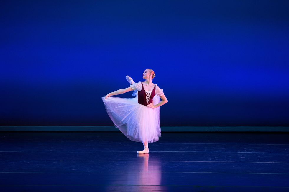 A blonde teenage ballet student in a long white tutu and burgundy peasant bodice performs onstage in front of a blue backdrop. Holding her skirt, she brushes her left leg into an attitude derriere while in pliu00e9 on her right leg, and looks to her right with a confident smile.