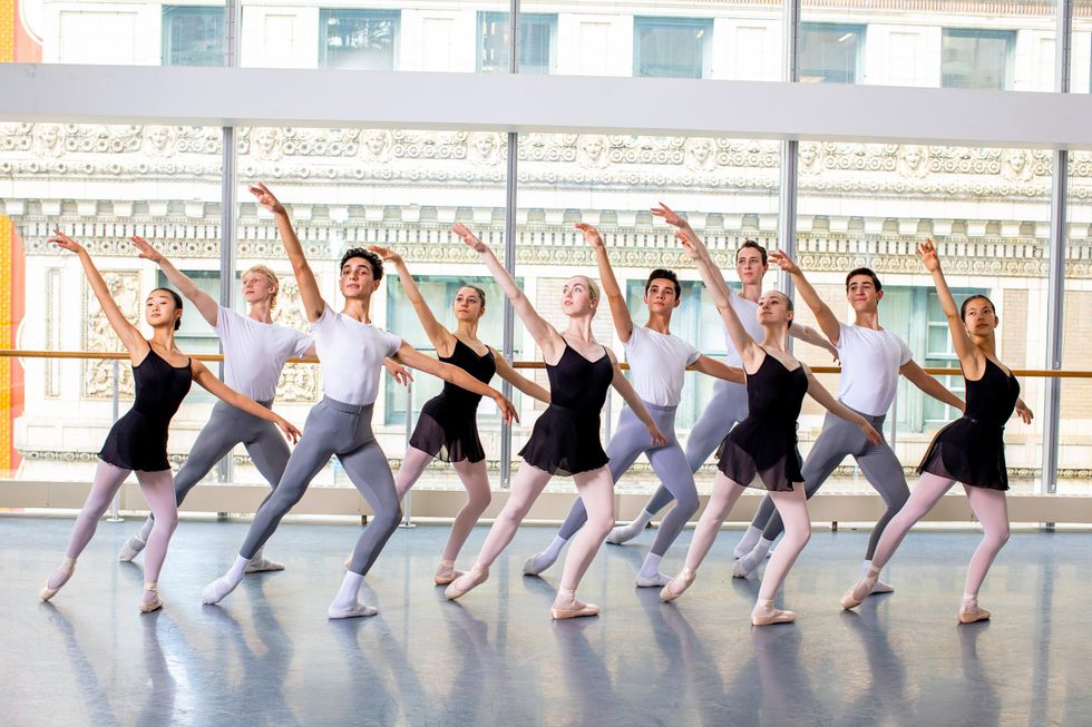 In front of large studio windows, 10 ballet studentsu2014five girls and five boysu2014stand in a staggered line and pose with their right leg in tendu derriere in effacu00e9. They all lift their right arm up high and look up towards their hand while their left arm is held low.