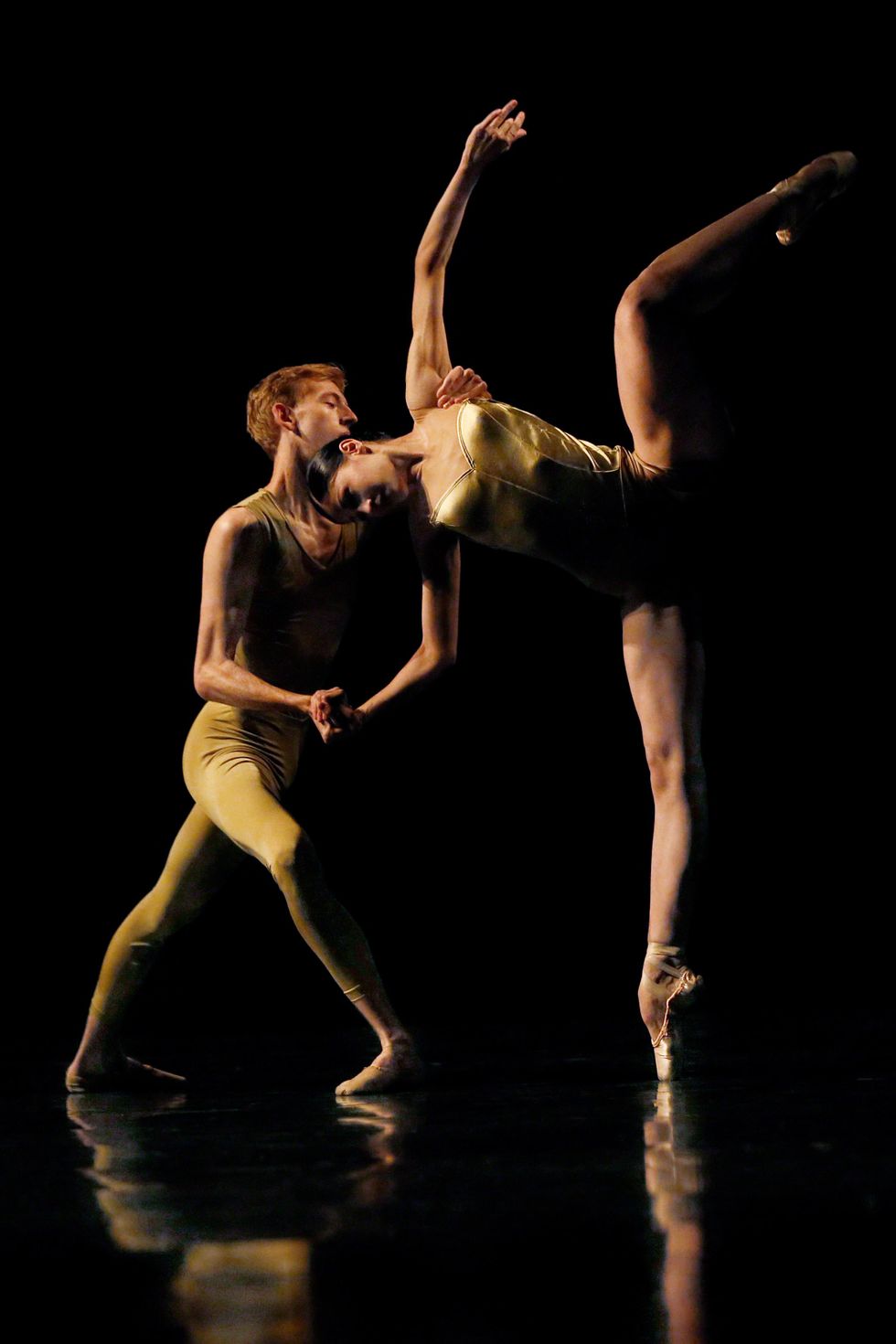 Wearing shimmery gold costumes, a male and female ballet student partner each other, lit from the left on a darkened stage. The boy lunges in croisu00e9 with his right foot in front and holds the girl's right hand and left should as she leans over in a side attitude.