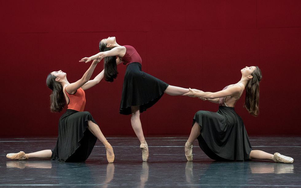 Three teenage ballet students pose onstage in profile, with the girl in the middle on pointe in degagu00e9 devant while reaching back with her upper body, and the two other dancers kneeling on each side of her. The dancer on her right holds her arms while the dancer on her left holds her right leg. They all wear long black skirts and leotards, and wear their long hair half pulled back.