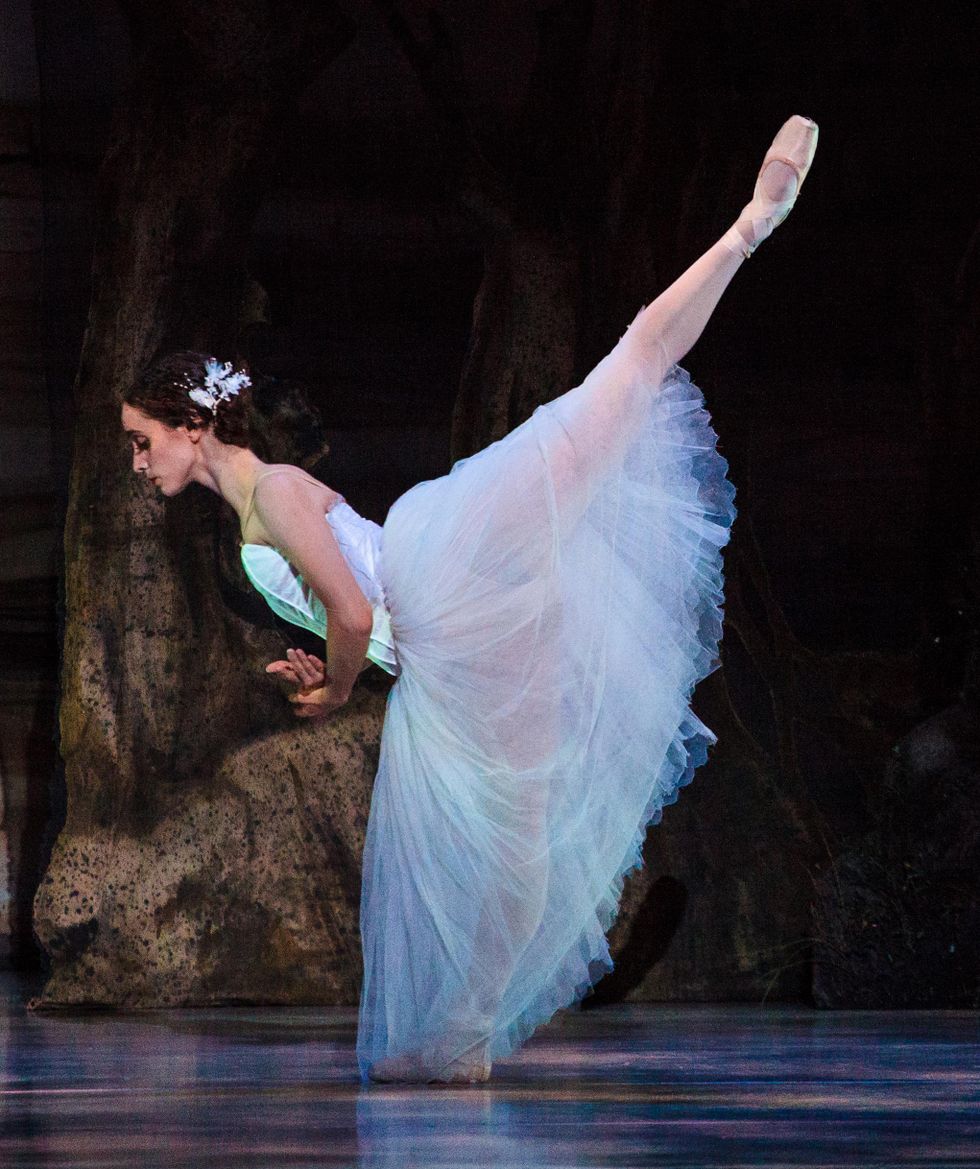 Arolyn Williams wears a long white Romantic tutu and pointe shoes and does a penchu00e9 on her right leg with her arms crossed in front of her chest. Behind her onstage is a wooded forest set.