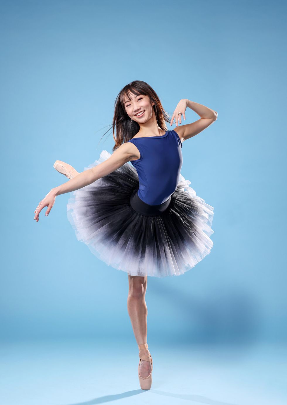 Chisako Oga, an Asian-American ballerina, poses in attitude derriere croisu00e9 on her right leg, with her right arm out to the side and her left hand grazing her left shoulder. She smiles happily towards the camera, her black hair blowing in the breeze, and wears a blue leotard, black-and-white ombre tutu, and skin-colored pointe shoes.