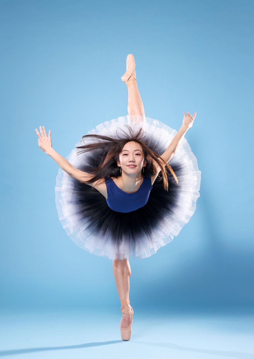 Chisako Oga, an Asian-American ballerina, does a penchu00e9 on pointe towards the camera with her arms held out to the side and her long hair flying. Smiling confidently, she wears a blue leotard and a black and white ombru00e9 tutu.