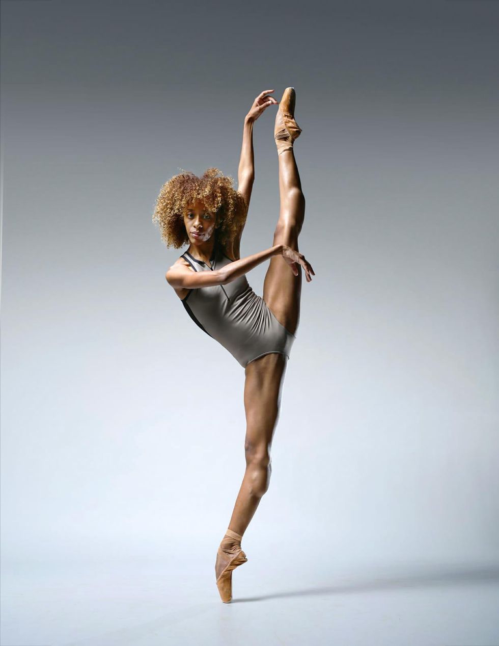 Chlou00e9 Lopes Gomes wears a gray leotard and tan pointe shoes and kicks her left leg high in the air. She extends her left arm up and her right arm across her chest, and looks confidently towards the camera.