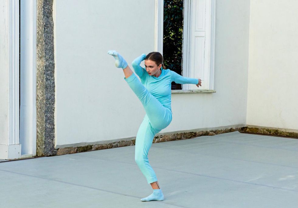 Jessy Dick, wearing a light blue turtleneck, pants and socks, kicks her left leg high and holds the side of her head with her right hand. Her right elbow is bent in towards her leg and she dances outdoors on a marley floor and in front of a white wall..