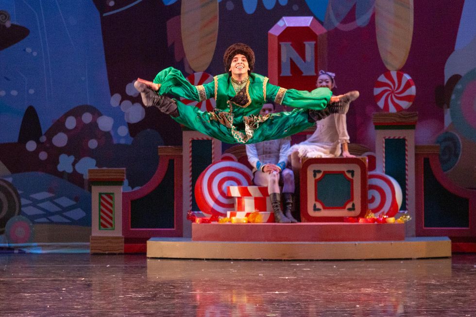 In front of a Kingdom of Sweets backdrop ,Alejandro Gonzu00e1lez performs a split jump in second position during a performance of The Nutcracker, his arms reaching for his feet. He wears loose-fitting pants and blouse made of shiny green fabric, a Russian fur hat and black boots.