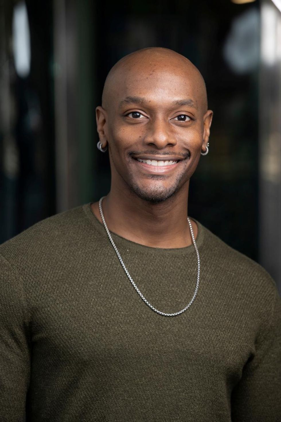 Jason Ambrose, shown from the chest up, smiles towards the camera. He wears a hunter green shirt, silver necklace and silver hoop earrings.