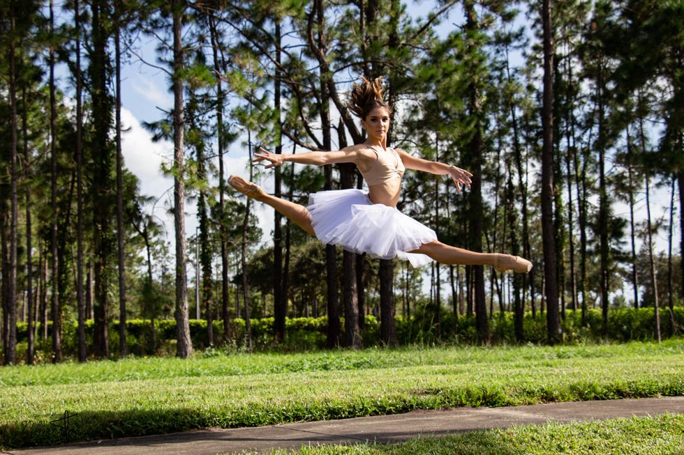 Wearing a white practice tutu, beige bralet and pointe shoes, Ximena Azurmendi performs a large split sissone outside in front of a grove of pine trees.