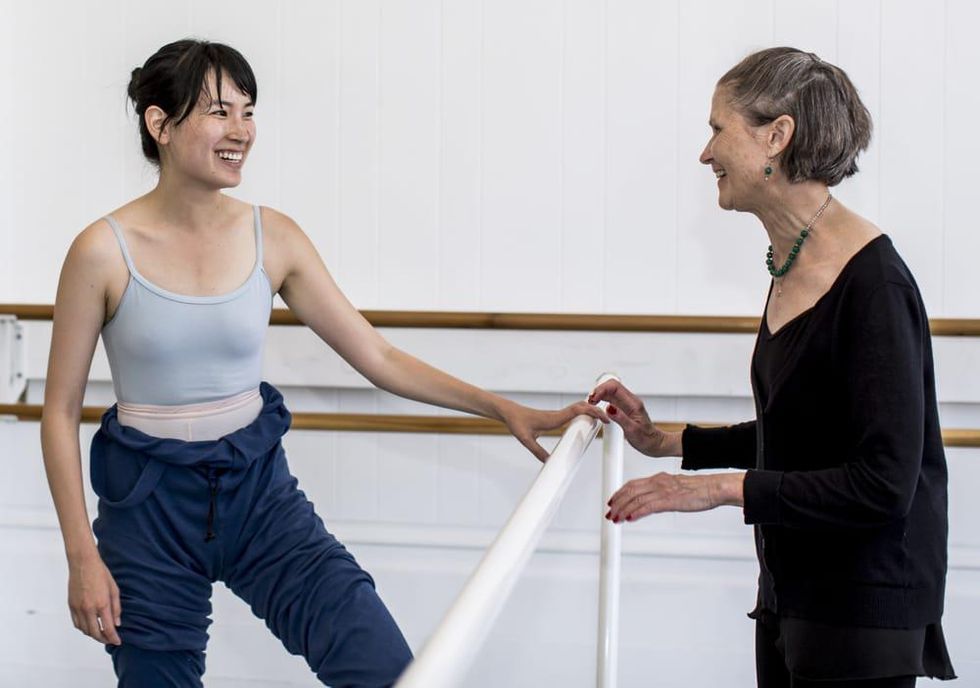 Kathy Mata, in a black sweater and ballet skirt, chats and smiles with one of her adult ballet students, who wears a leotard and jumper rolled down at the waist.  They stands across from each other with a white portable ballet barre in between them in a brightly lit dance studio.