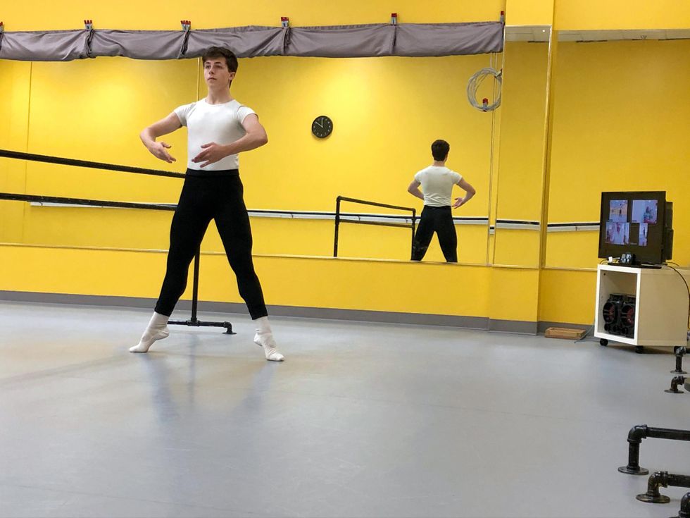 Cooper Sainiak, a white teenage ballet student, balances in second position relevu00e9 at the barre with his arms in first position. He wears a white T-shirt, black tights and white socks and slippers, and dances in a dance studio with bright yellow walls. A TV showing a virtual Zoom class is in the background.