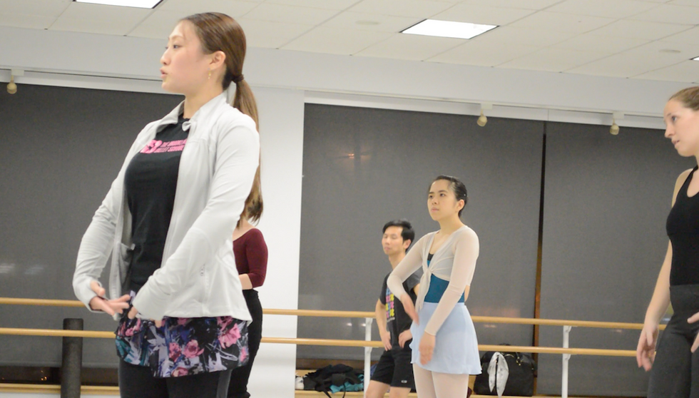Haruka Tamura stands at the front of her class with her arms in fifth position en bas in a dance studio while a group of adult students observe her from behind. She wears a floral ballet skirt, black T-shirt and gray zip-up sweater, while the dancers wear various dancewear.