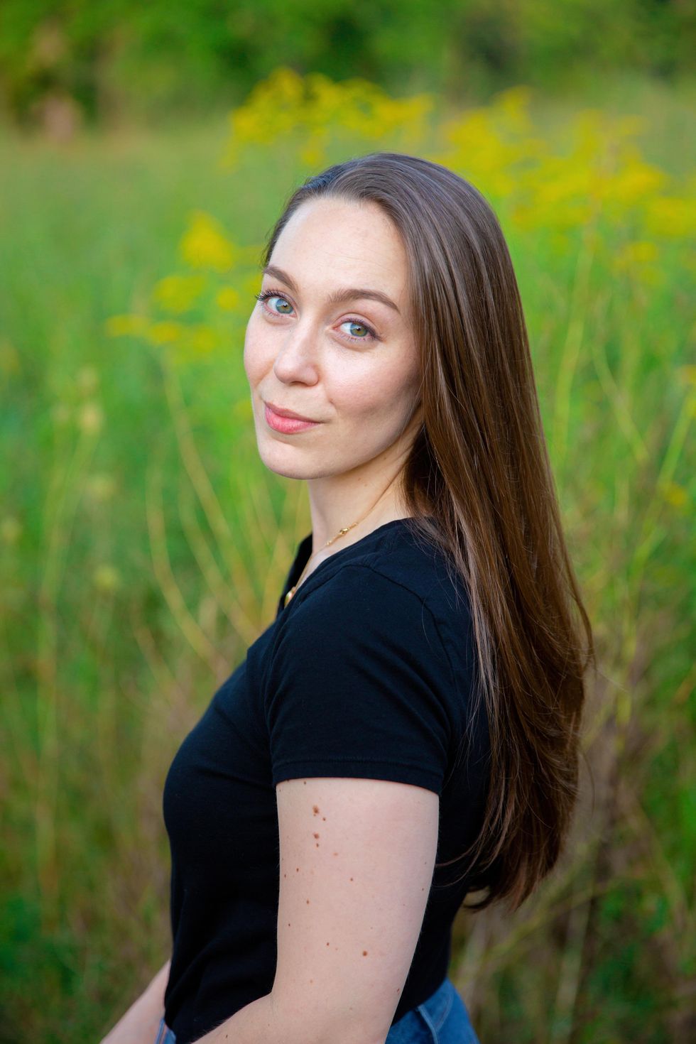 Chloe Angyal, wearing a black short-sleeved T-shirt, is shown from the waist up in a field, looking over her shoulder and smiling serenely towards the camera. Her long brown hair flows down her back.