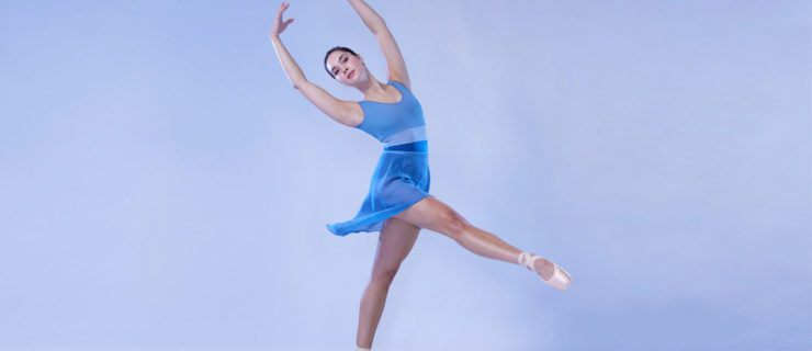 Mira Nadon, wearing a blue leotard and ballet skirt and pink pointe shoes, stands on pointe on her left foot and reaches her right leg forward in degagé. She twists her upper body towatrds the camera and holds her arms in high fifth position.