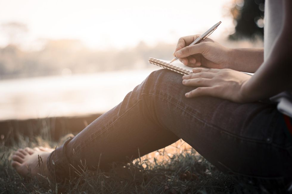 A barefoot woman in jeans sits in the grass and writes in a notebook.