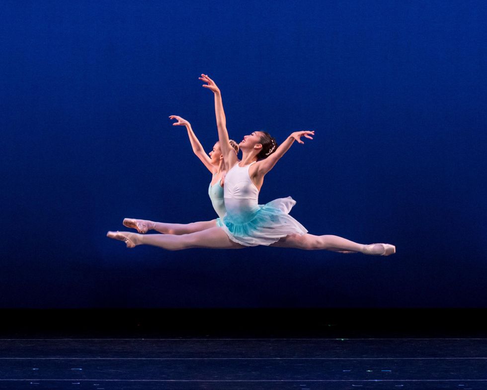 Melissa Meg and Madelaine Boyce, two female ballet dancers, perform a grand jetu00e9 towards stage right in front of a bright blue backdrop. Each wears a white and green dance dress, pink tights and pink pointe shoes.