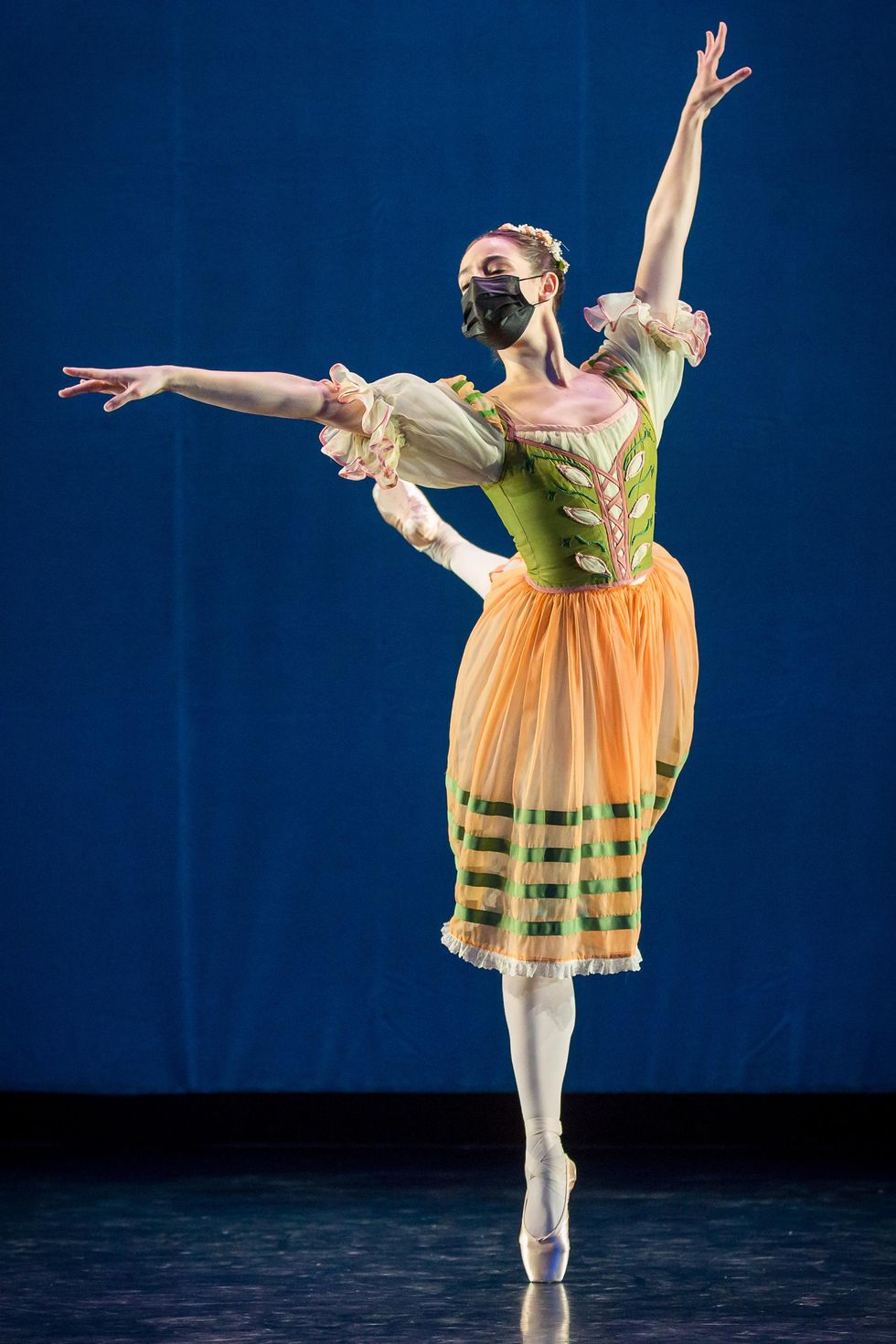 Itzel Hernandez does an attitude croisu00e9 balancing on her right leg in front of a blue backdrop. She wears a green and oragne peasant dress, pink tights, pointe shoes and a black face mask.