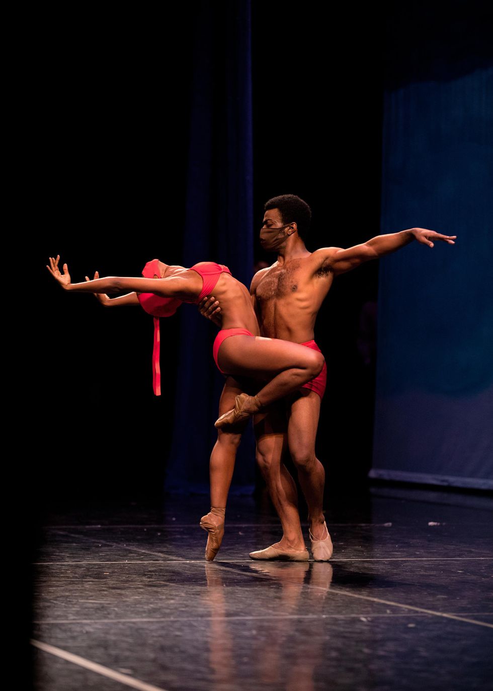 On a darkened stage, Paunika Jones balances in passu00e9 and does cambru00e9 back while her partner, WIlliam Moore, Jr., holds her waist with his right hand and extends his left arm in second. She wears a red bikini, face mask and pointe shoes while he wears red shorts and brown ballet slippers.