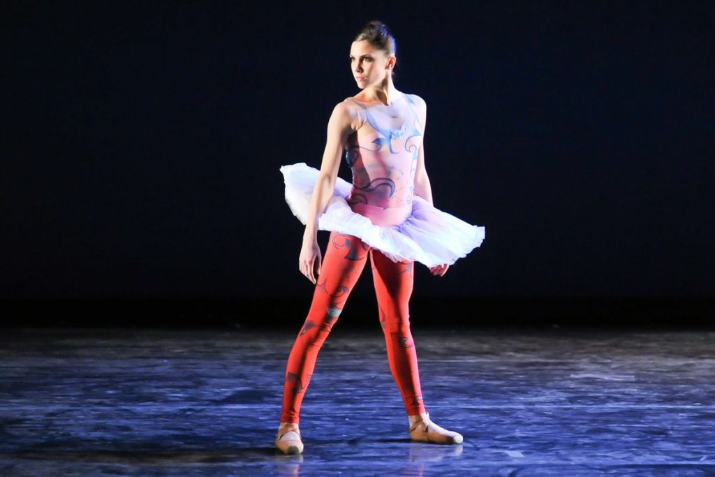 Caitlin Peabody stands ina. parallel second position onstage and looks out over her right shoulder. She wears an ombre pink and red unitard with a swirly pattern and pancake tutu.