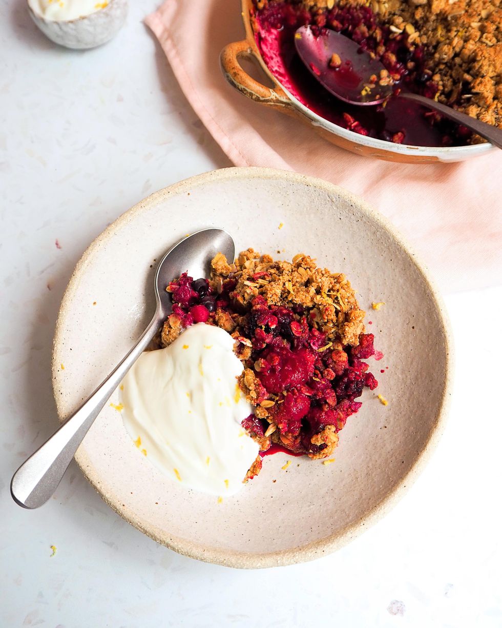 A rustic ceramic bowl with plain yogurt, a spoon and a berry crisp inside is photographed from above.