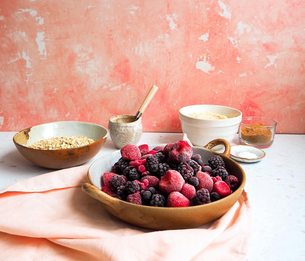 A ceramic bowl containing frozen strawberries, raspberries and blueberries sits on top of a loose, pink cloth napkin. Bowls of oats, sugar and flour rest behind the berries, all on a white tabletop and in front of a pink sponge-painted background.