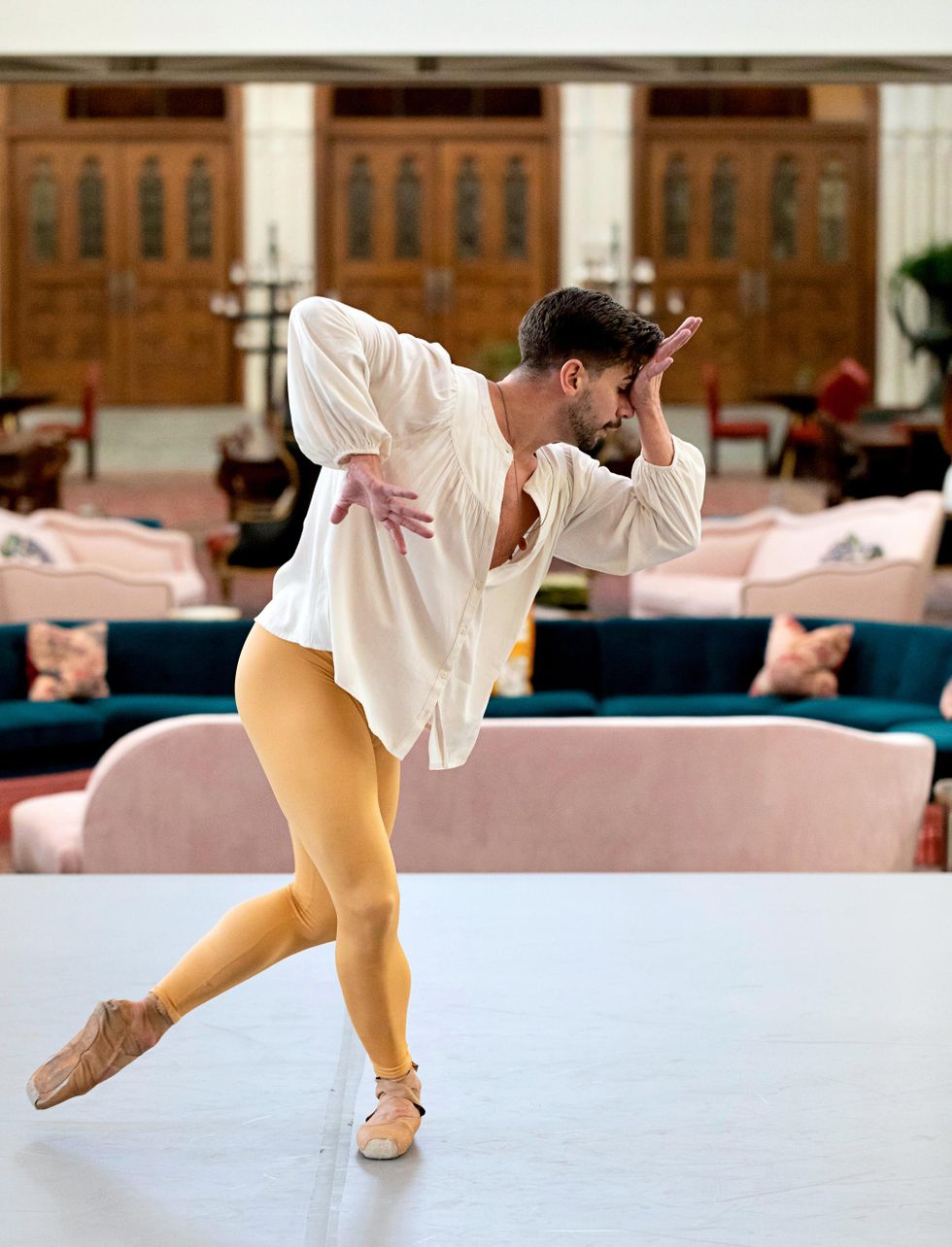 Brian Gephart poses in B-plus and bends forward with his left hand to his forehead and his right hand pushing downward. He wears yellow leggings, pointe shoes and a white peasant blouse.