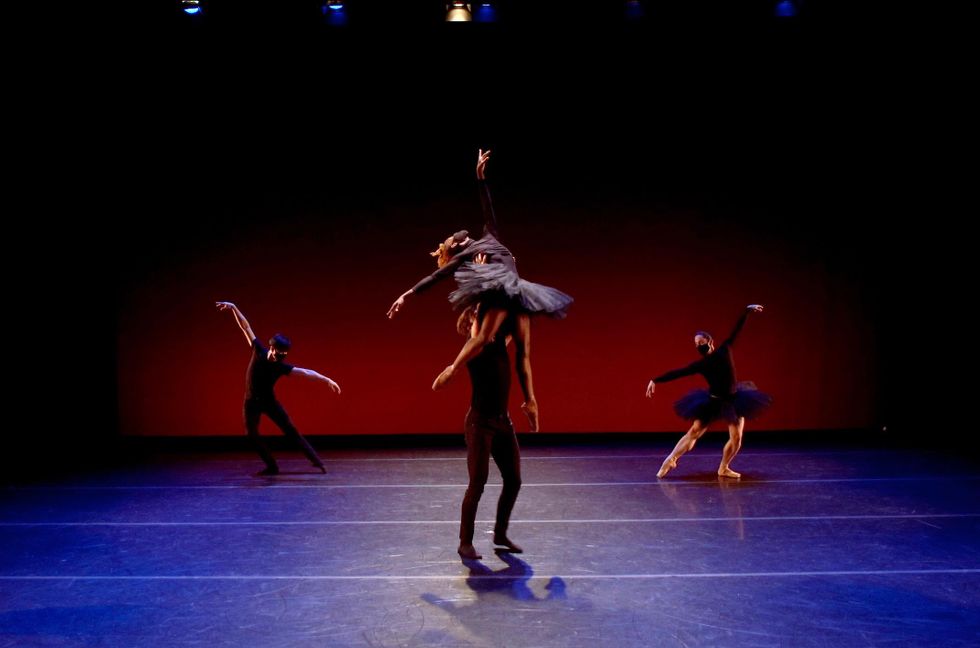 A male dancer in all black lifts another male dancer in a gray tutu and pointe shoes above his head. Behind them on either side are two other male dancers lunging out in tendu second. The dancer on the right wears a black tutu, and pointe shoes while the dancer on the left wears black tights, ballet slippers and a black T-shirt. All of the dancers wear face masks.