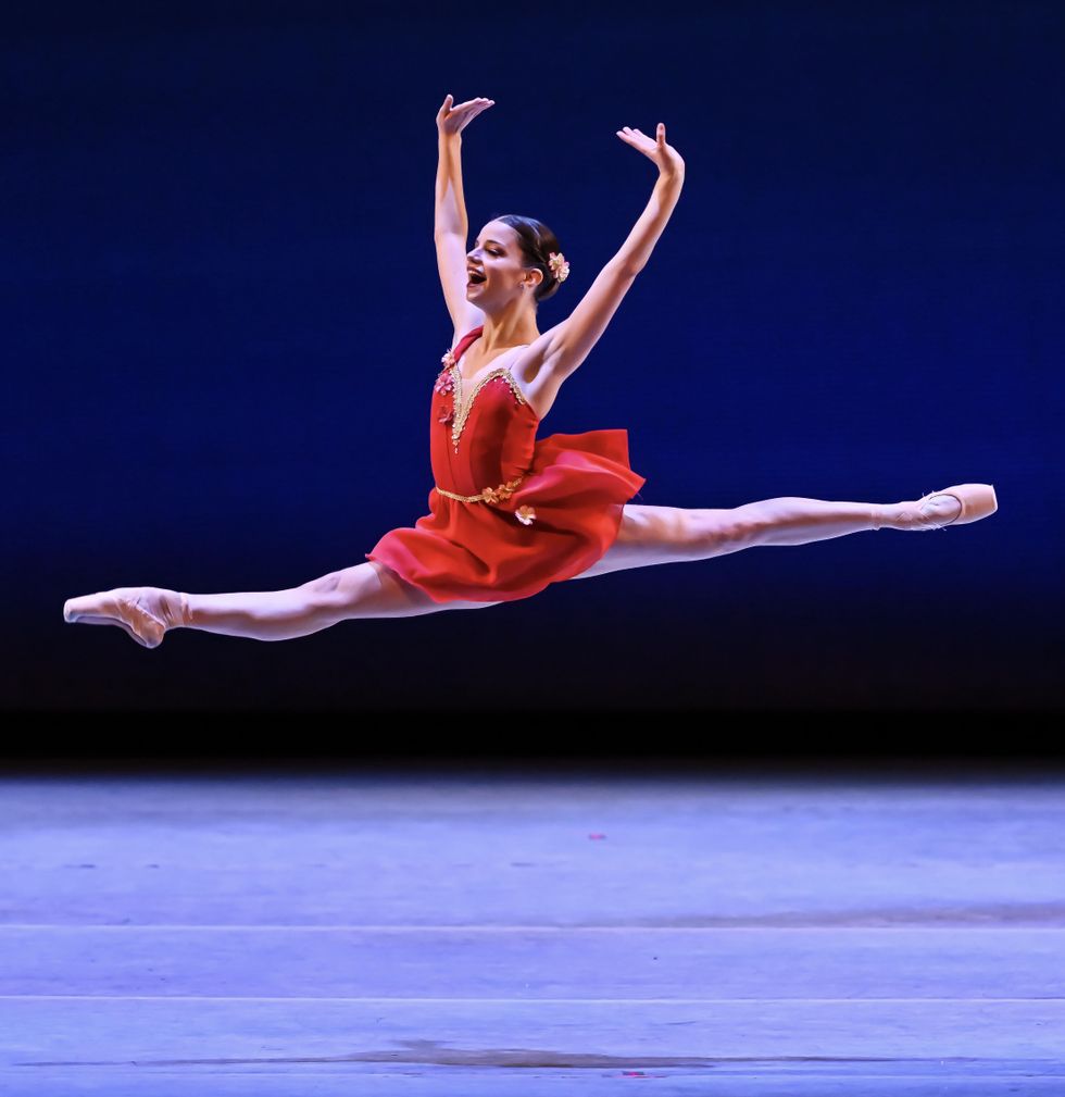 Clara Thiele soars through the air in a grand jetu00e9 above the stage, mouth wide open, hands flexed above her head.