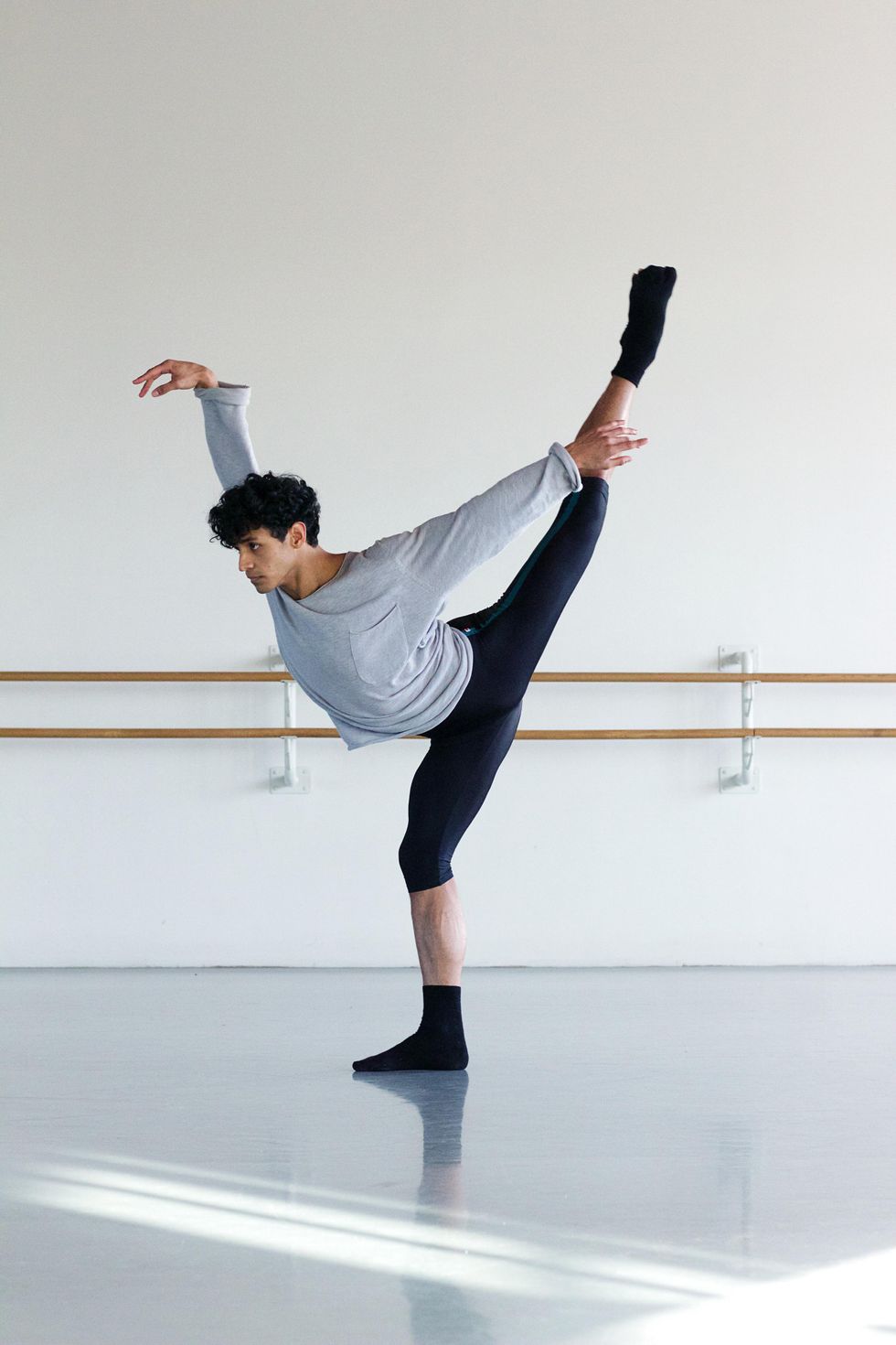 Ricardo Urbina Reyes, wearing a relaxed gray T-shirt, black knee-length biker pants and black socks and ballet slippers, stands on his right leg in pliu00e9 and kicks his left leg and right arm high in first arabesque.