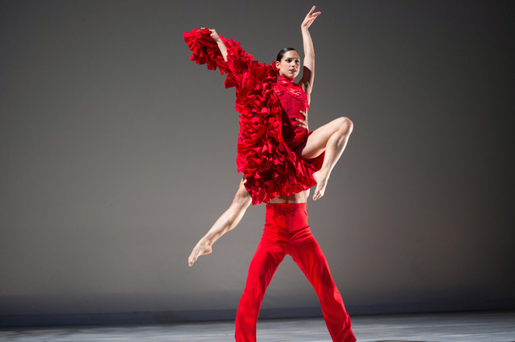 Melissa Verdecia wears a dramatic red costume with a long, ruffled skirt. She holds the skirt up with her right hand and jumps up while her partner, wearing red pants, lifts her from behind.