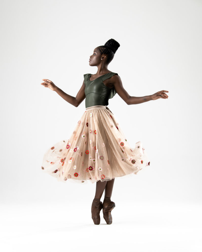 Ingrid Silva stands in fifth position en pointe. She wears a dark green leotard underneath a knee-length pink skirt embroidered with red and white flowers. The skirt is flowing in the air, as if she just dropped it, and her arms are floating at her sides. 