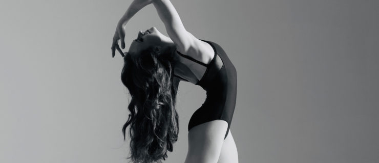 In this black and white ballet photo, Alexandra Martin stands in fourth position and arches her head back, her long hair flowing behind her. She wears a dark leotard and reaches her right hand back towards her forehead, her eyes closed.