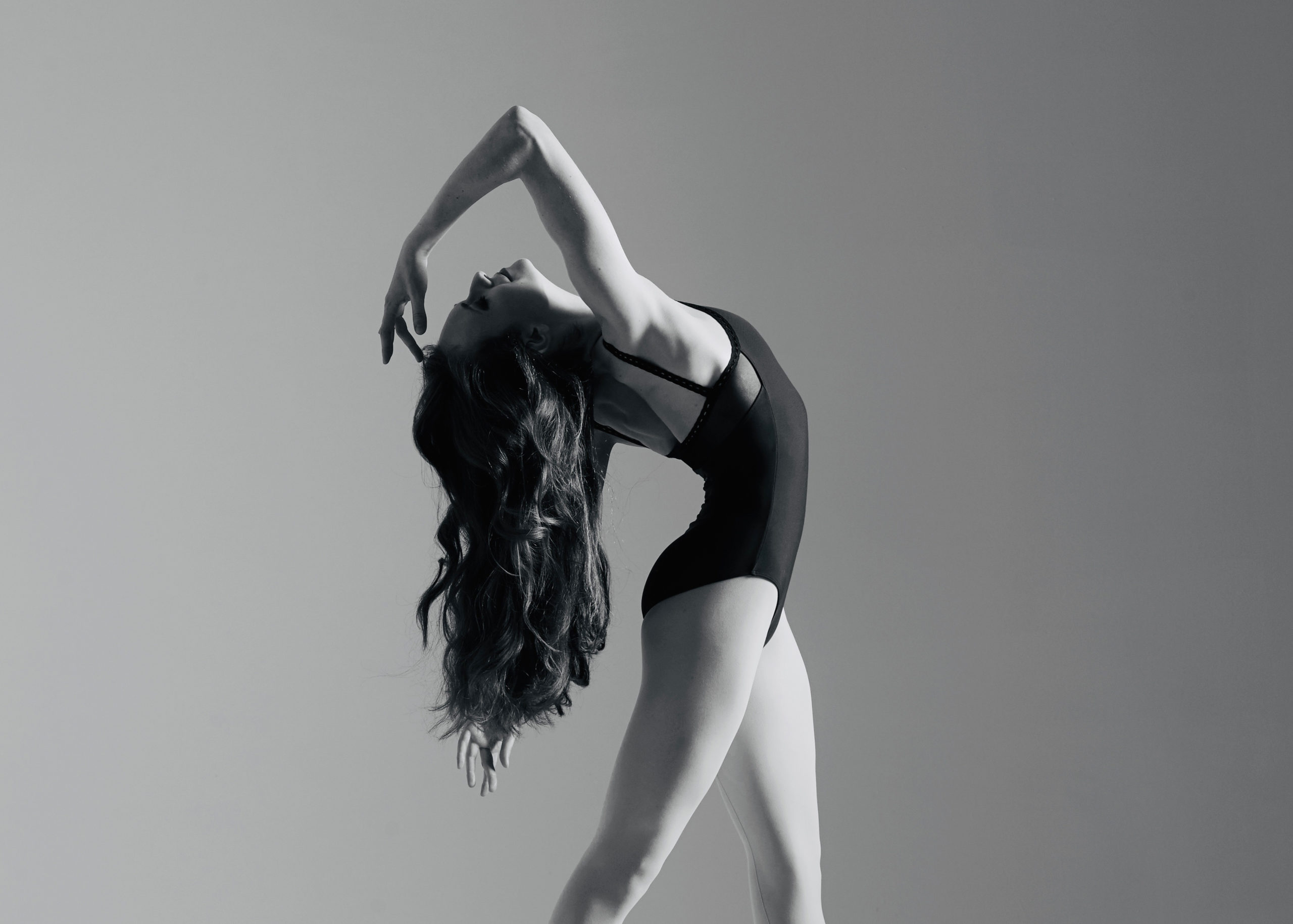 In this black and white ballet photo, Alexandra Martin stands in fourth position and arches her head back, her long hair flowing behind her. She wears a dark leotard and reaches her right hand back towards her forehead, her eyes closed.