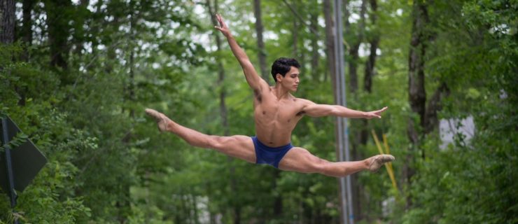 On a wooded street, Andrei Chagas does a large split jump in second with his right arm lifted slightly. He wears blue booty shorts and looks out over his left arm, which is out to the side.