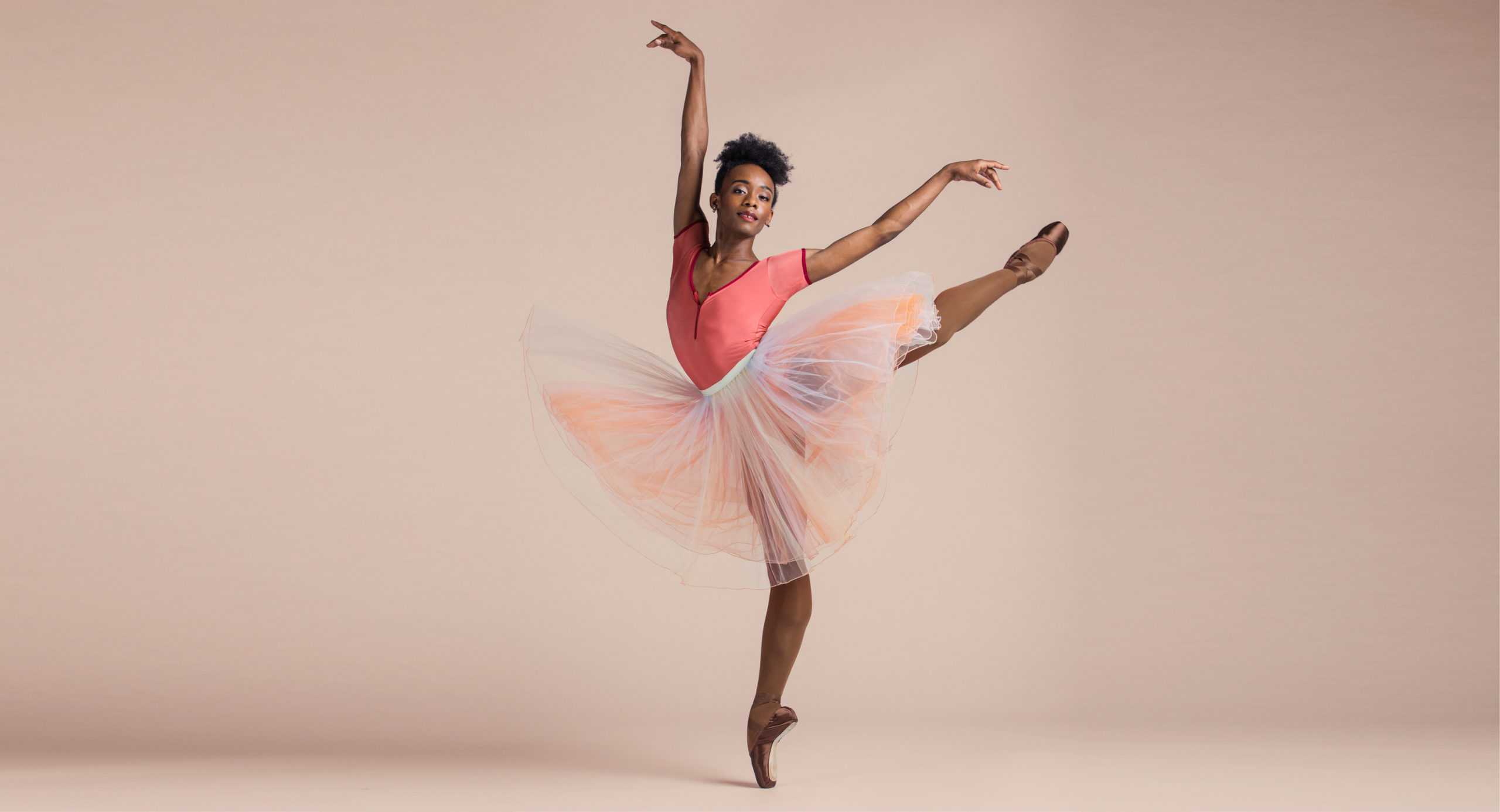 Ashton Edwards wears a pink leotard, brown pointe shoes and a long, peach tutu. He stands in front of a taupe backdrop and performs a pique first arabesque on his right leg and looks confidently towards the camera.