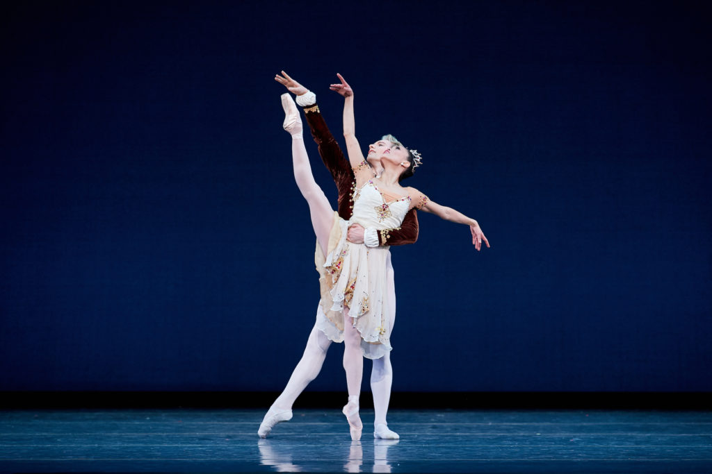 Patrick Yocum stands behind Lia Cirio onstage and holds her around the waist with his left arm as she performs a développé ecarté on pointe with her right leg. Cirio is costumed in an off-white camisole dress with floral embellishments and a floral headpiece; Yocum wears white tights and a brown velvet jacket.