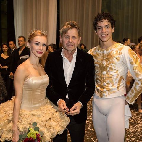 Mikhail Baryshnikov, wearing a sport coat and black pants, stands between Iana Salenko and Issac Hernández backstage at a theater and pose for a picture. Salenko wears an ivory tutu while Hernandez wears white tights and a white and gold jacket.
