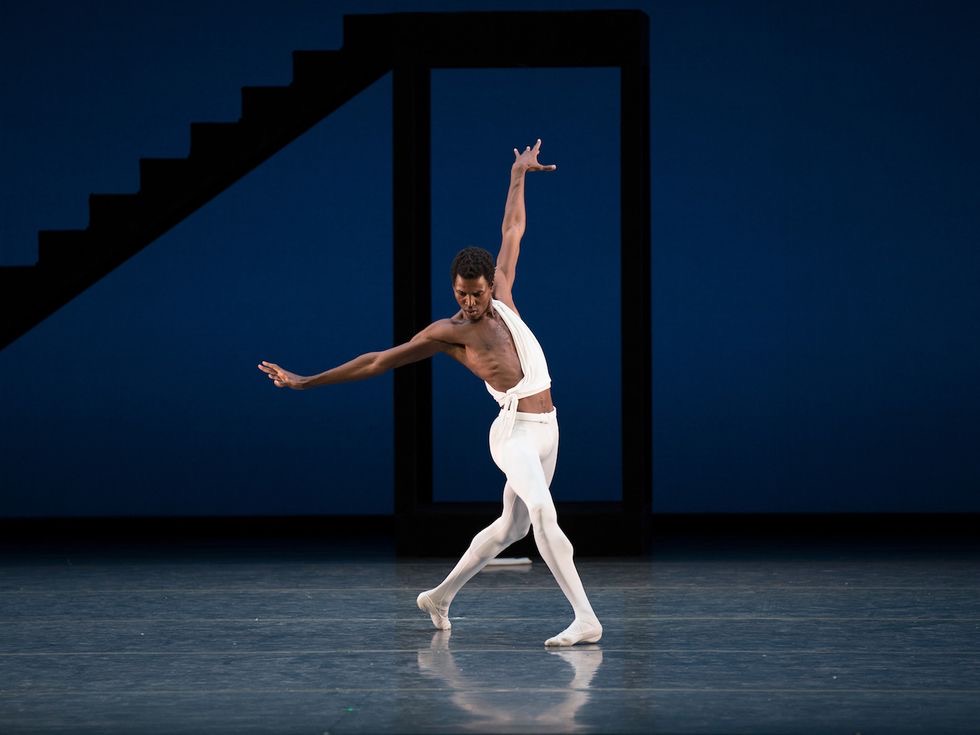 Calvin Royal III, wearing a white Grecian tunic and white tights and slippers, steps forward on his right leg in croisé with his arms out to the side. He looks down at his feet, as if trying to catch his balance. Behind him onstage is a tall, stark black staircase and a blue backdrop.
