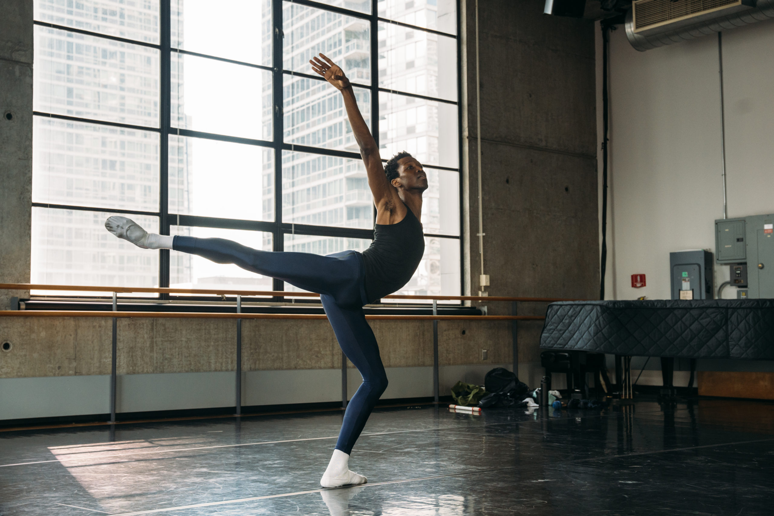 In a dance studio with large, industrial windows, Calvin Royal III performs an arabesque allongé with his right leg and arm raised. He wears blue tights, a dark tank top and white socks and ballet slippers, and looks out towards the front of the room.