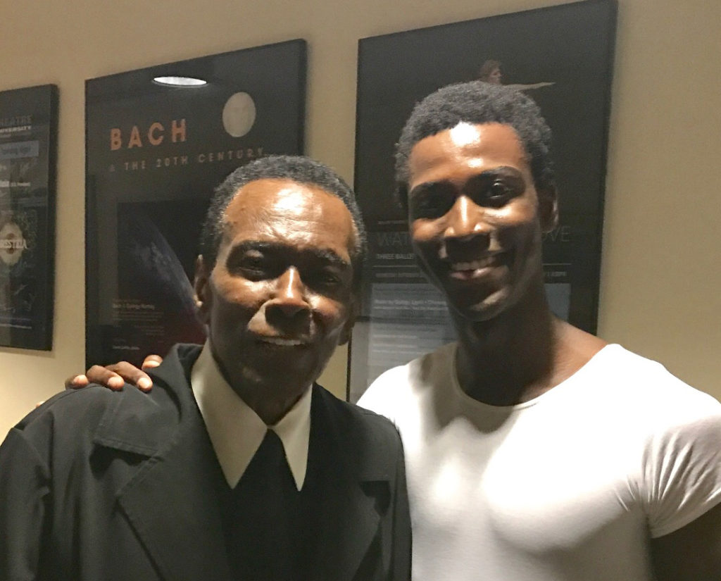 Arthur Mitchell, in a black suit coat, tie and white shirt, stands next to Calvin Royal III with his arm around his waist. Royal, in a white T-shirt and black tights, has his right hand around Mitchell's shoulder, and they both smile for the camera.