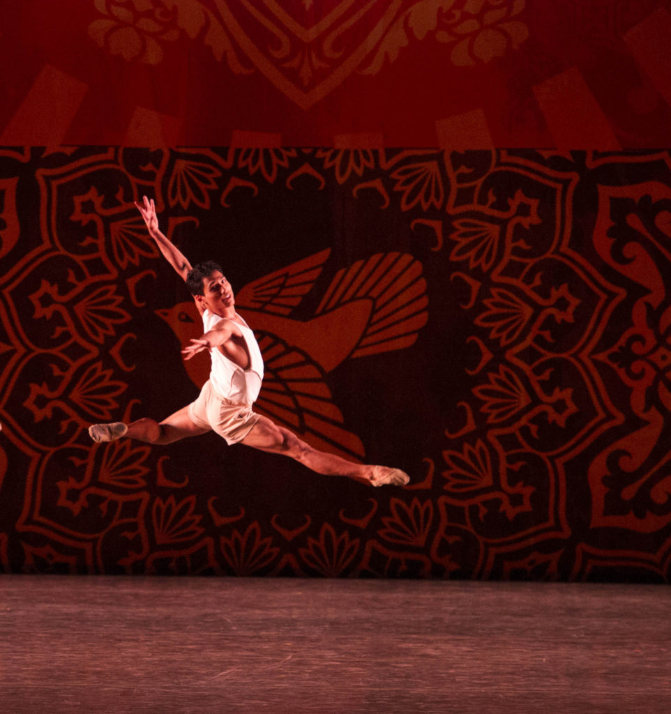 Andrei Chagas leaps with his right leg in front and his left leg bent behind him and looks out towards the audience. Behind him is a printed backdrop with an illustration of a dove and other designs. He wears a white shirt and beige shorts.