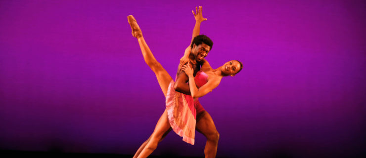 Amanda Smith, in a pink dance dress, leans back into a développé devant while wrapping her arms around Da'Von Doane's shoulders. He holds her around the waist and lunges on his left leg. Both dancers look towards the audience and smile brightly.