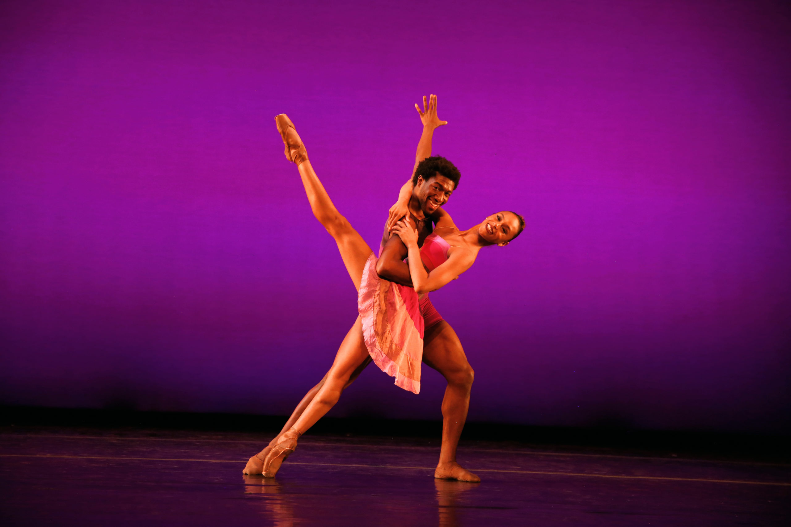 Amanda Smith, in a pink dance dress, leans back into a développé devant while wrapping her arms around Da'Von Doane's shoulders. He holds her around the waist and lunges on his left leg. Both dancers look towards the audience and smile brightly.
