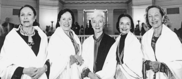 In this black and white photo, Maria Tallchief, Marjorie Tallchief, Rosella Hightower, Moscelyne Larkin and Yvonne Chouteu stand close together in a line and smile towards the camera. They are standing in a grand, open foyer and wear identical Native American shawls .