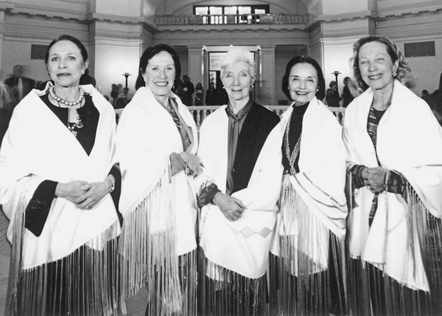 In this black and white photo, Maria Tallchief, Marjorie Tallchief, Rosella Hightower, Moscelyne Larkin and Yvonne Chouteu stand close together in a line and smile towards the camera. They are standing in a grand, open foyer and wear identical Native American shawls .