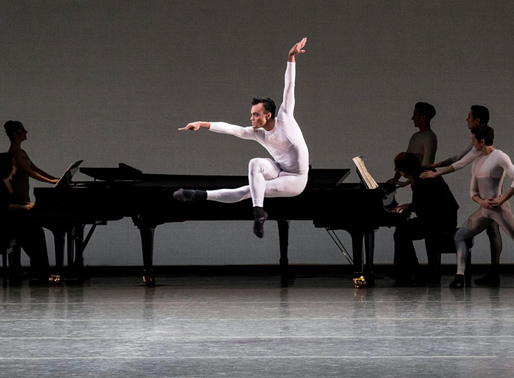 Taylor Stanley, wearing a white unitard, jumps up with his left leg straight out and his right leg bending over it. He reaches his right arm out in front of him and looks over it while lifting his left arm up above his head. Behind him onstage are two grand pianos and musicians and dancers in silhouette.