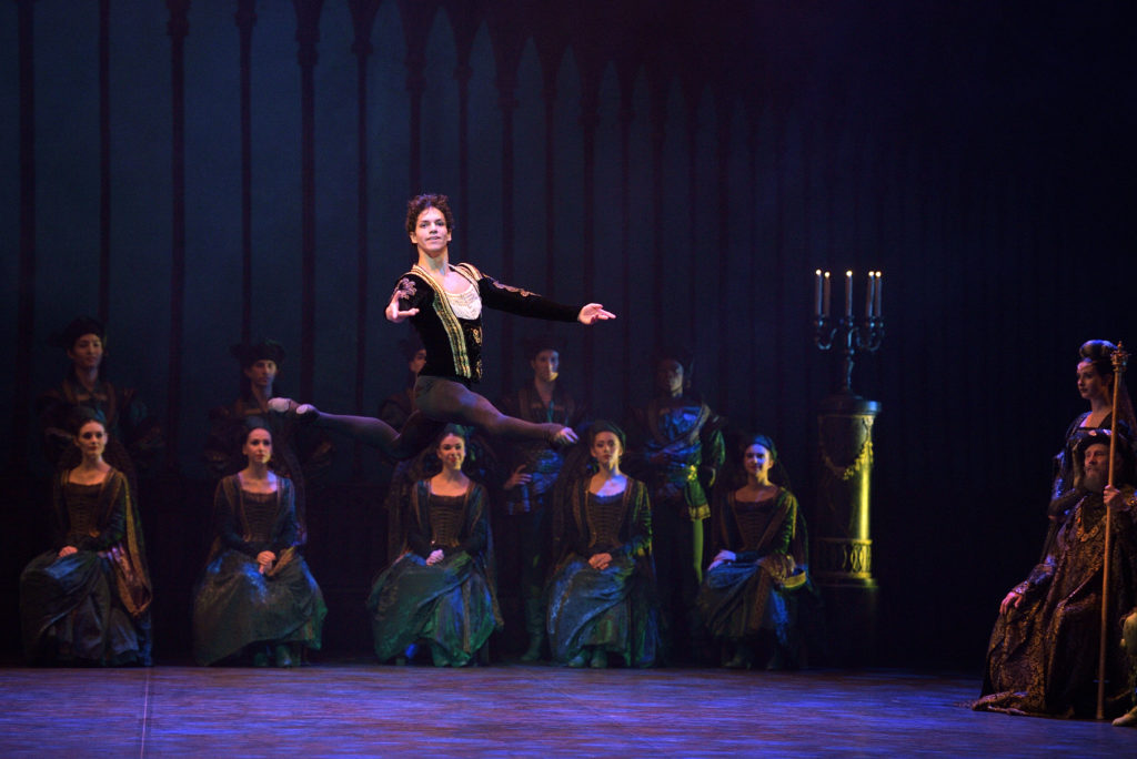 Issac Hernández performs a grand jeté en avant with his left leg in attitude during a performance of Swan Lake. He weara black tights and a black velvet tunic, and behind him onstage sits a row of aristocratically costumed women and men.