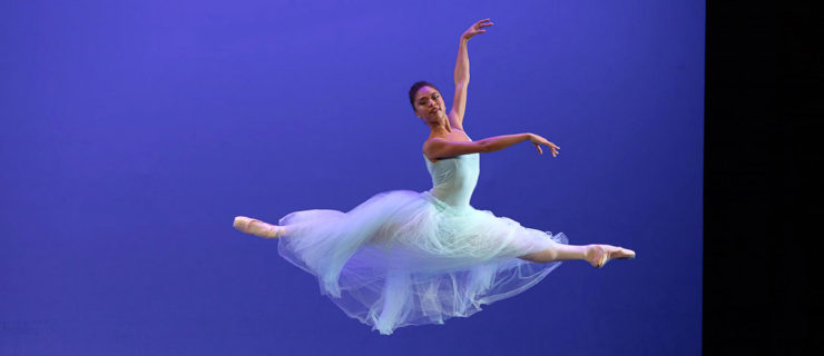Jasmine Perry wears a light blue, ankle-length tutu and performs a saut de chat to the left with her arms in third arabesque.