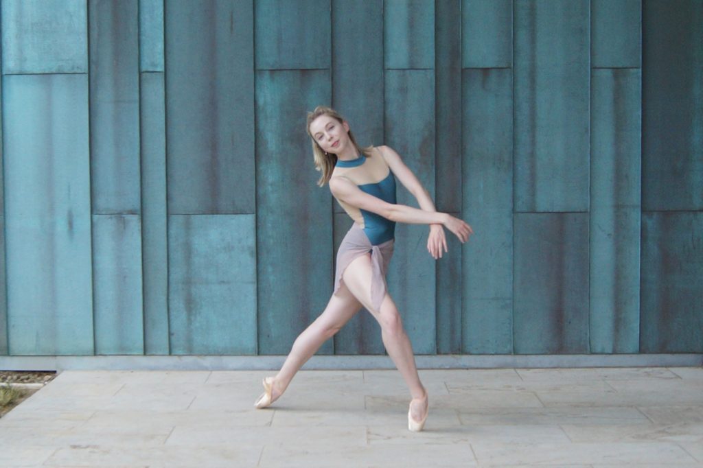 Camille Kellems balances in a lunge on pointe, with her right leg in front and bent and her left leg extended straight behind her. She crosses her wrists and leans slightly to the right and looks towards the camera. She wears a blue leotard and grey skirt.